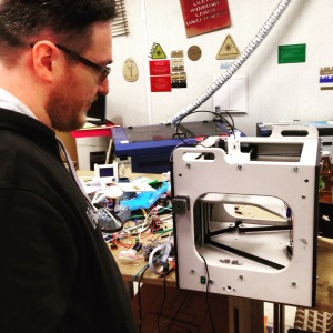 A man standing beside a small personal scale 3D printer, sitting on a desk with a lot of wires and other crafting tools. 